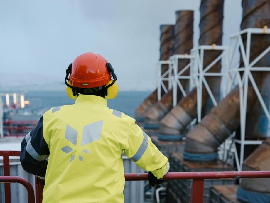A person wearing a red safety helmet and a high-visibility yellow jacket with the Equinor logo stands with their back turned, looking out over the gas facility at Melkøya. The sea can be glimpsed in the background.