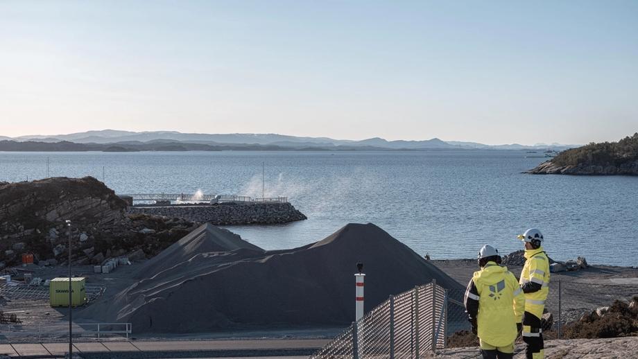 To personer ikledd refleksgult arbeidstøy med Equinor-logo på ryggen og hvit vernehjelm, ser utover Northern Lights-anlegget som oppføres ved sjøen i Øygarden. 