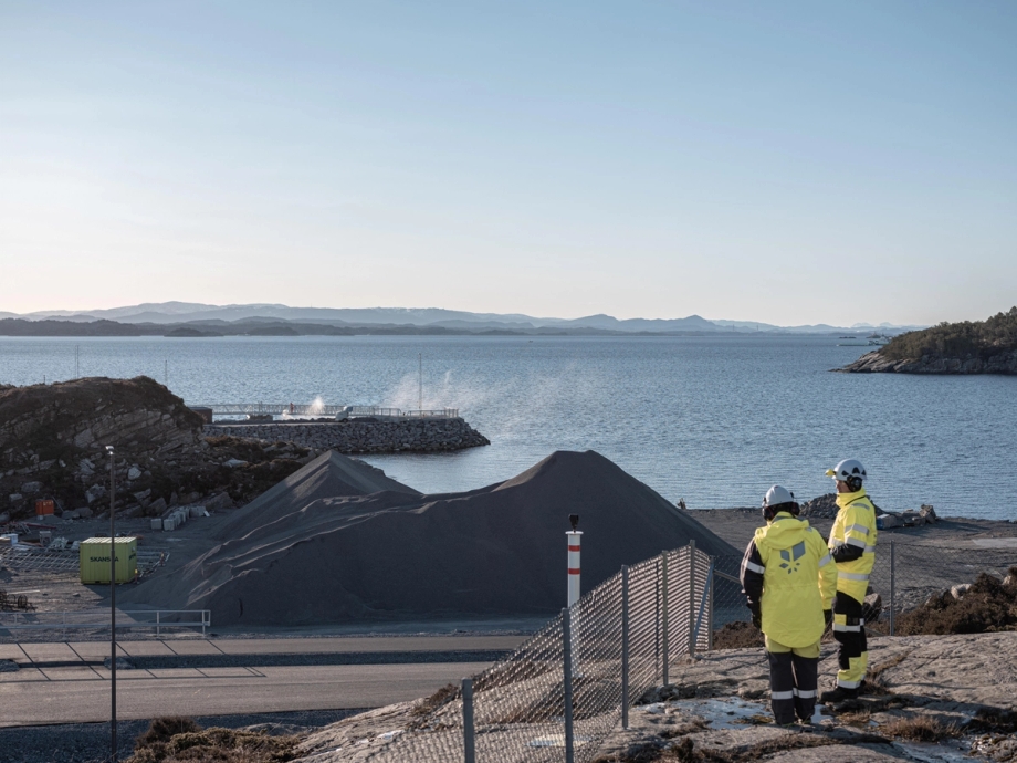 Two people wearing reflective yellow work clothes with the Equinor logo on the back and white safety helmets, look out over the Northern Lights facility being built by the sea in Øygarden. 