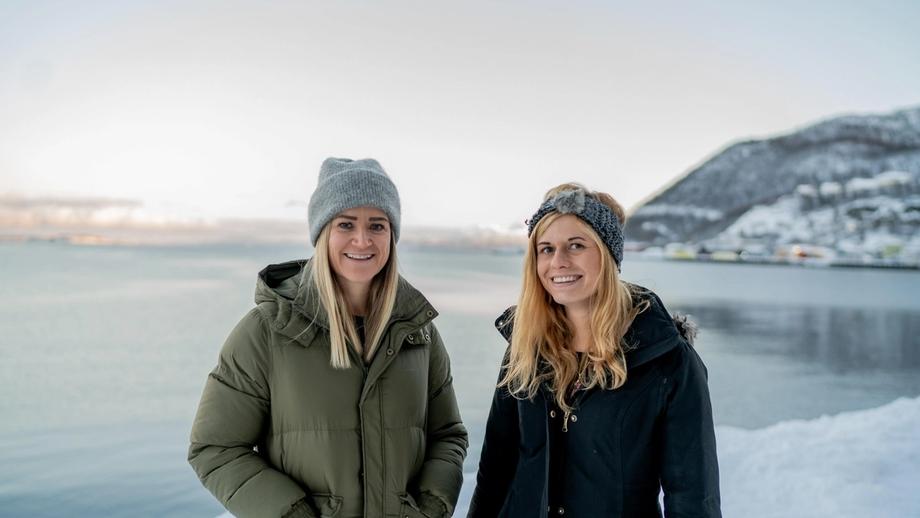 Portrettfoto av Linn Kristensen og Zoë Cumberpatch ved Equinor i Harstad som står utendørs i vinterklær, og med hav og snødekte fjell i bakgrunnen.