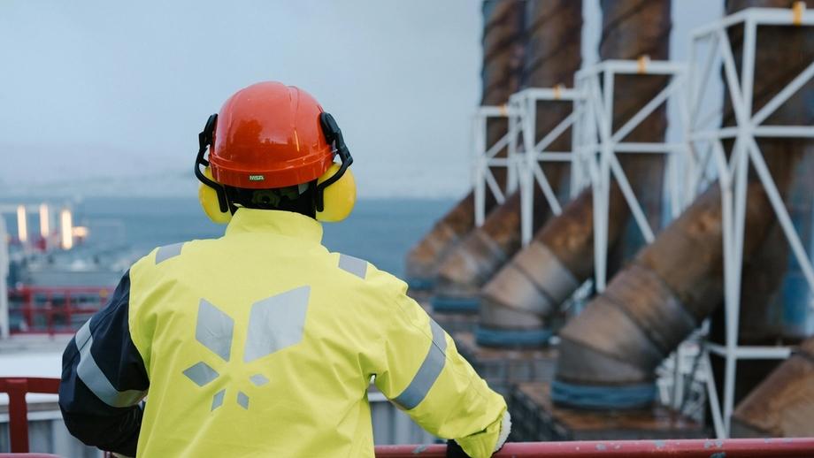 A person wearing a red safety helmet and a high-visibility yellow jacket with the Equinor logo stands with their back turned, looking out over the gas facility at Melkøya. The sea can be glimpsed in the background.